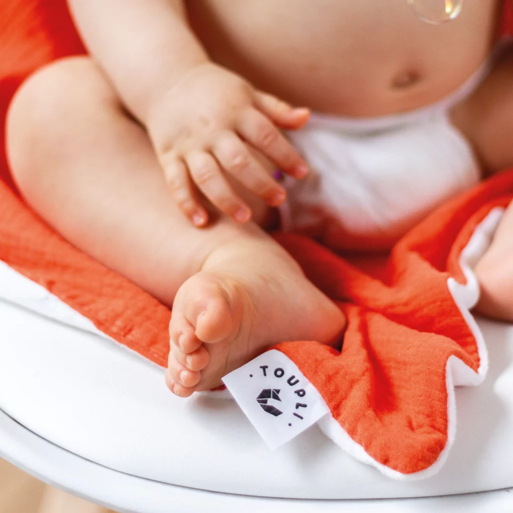 Gros plan sur un bébé assis sur un plaid orange Toupli